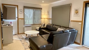 A living room with a black leather sofa, a white coffee table, and a patterned rug. There is a window with blinds, a lamp, and a door leading to another room. - Maddison-Chalet (Dinner Plain)