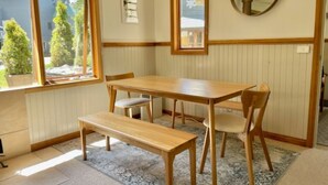 A dining area with a wooden table and benches, a round mirror on the wall, and a window with a view of trees and a house. - Maddison-Chalet (Dinner Plain)