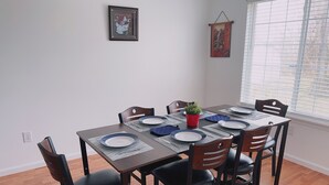 Un coin repas avec une table en bois dressée pour six personnes, entourée de chaises. Un tableau encadré orne le mur, et une fenêtre est dotée de stores.