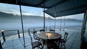 Ein überdachter Sitzbereich im Freien mit einem runden Tisch und Stühlen, mit Blick auf ein Gewässer und eine neblige Landschaft im Hintergrund.