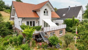 Une maison à deux étages, avec un toit recouvert de tuiles rouges, un balcon et un jardin où poussent différentes plantes et fleurs.
