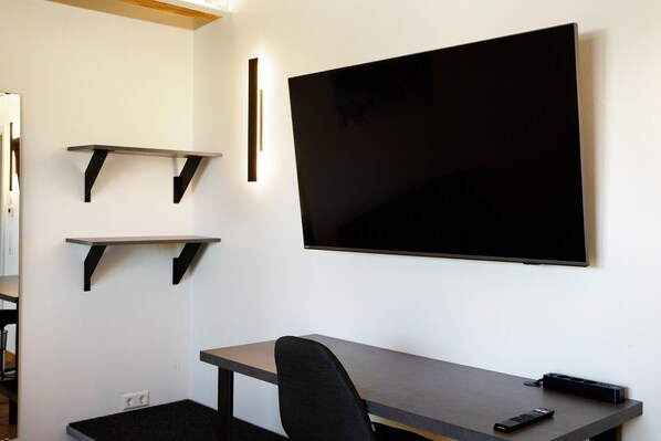 Ein Zimmer mit flachbildfernseher an der Wand, einem Schreibtisch mit Stuhl und zwei Regalen an der Wand.