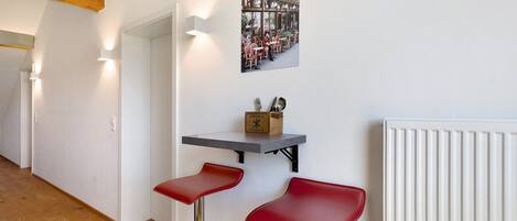 A hallway with a white wall, a radiator, a wooden floor, a table with a box, and two red bar stools.