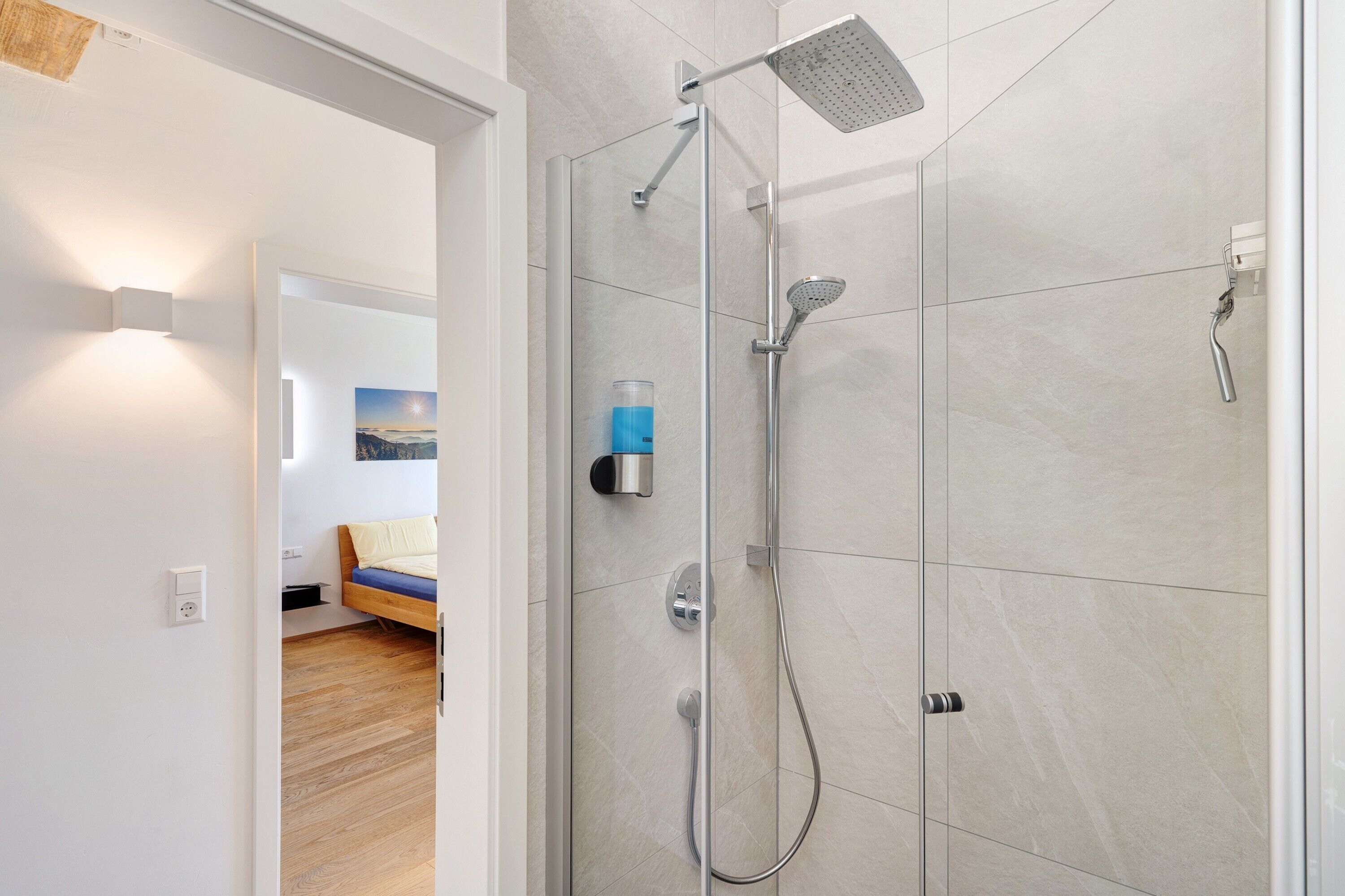 A modern shower with a handheld showerhead and a fixed overhead showerhead, a glass of water on the wall, and a bed with a blue mattress in the adjacent room.