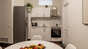 A modern kitchen with white cabinets, a stainless steel refrigerator, a microwave, and an oven. There is a plant on the countertop and a fruit bowl on the table.
