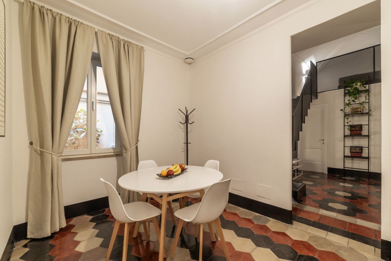 Un coin repas avec une table ronde, des chaises blanches et un saladier rempli de fruits. On aperçoit un porte-manteau et un escalier au fond.