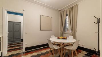 A dining area with a round table, white chairs, and a fruit bowl. A coat rack is visible on the right.