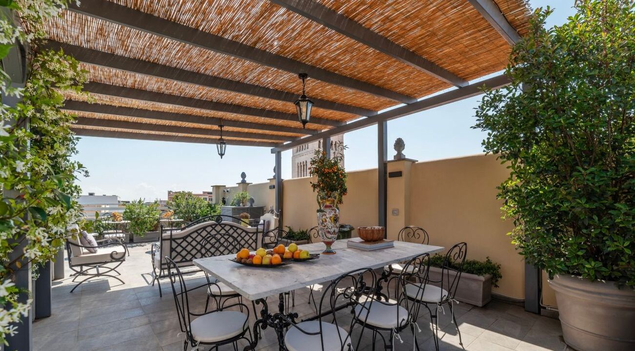 An outdoor patio with a table and chairs, surrounded by potted plants and a thatched roof.