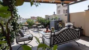A rooftop patio with outdoor furniture, potted plants, and a view of buildings in the background.