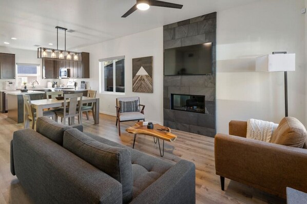 A modern living room with a fireplace, grey sofas, and a dining area with a kitchen in the background.