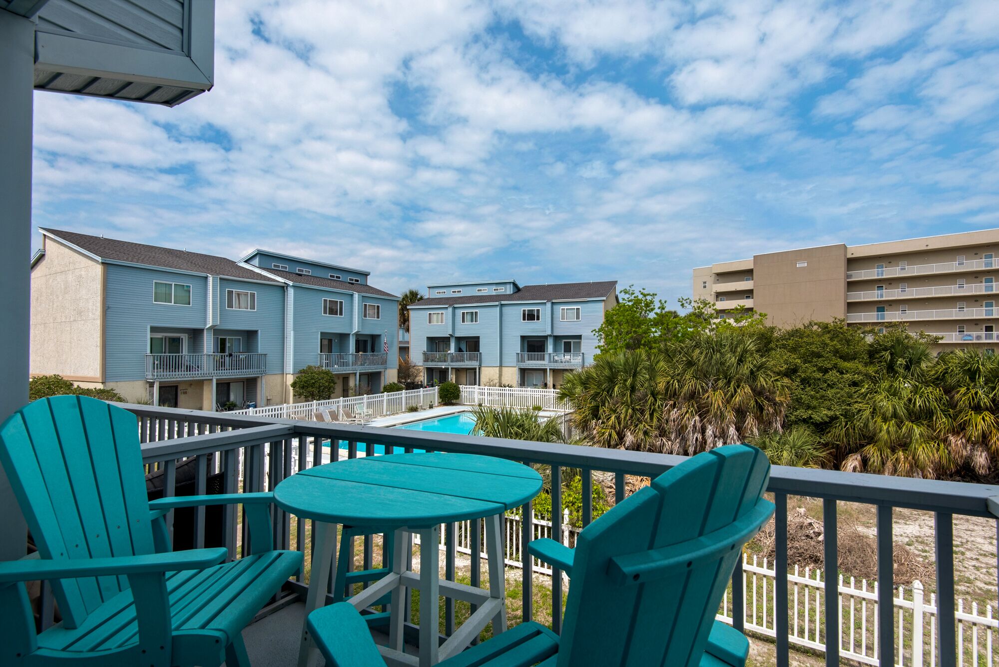 Un balcon avec des chaises turquoise et une petite table, donnant sur une piscine et les bâtiments avoisinants.
