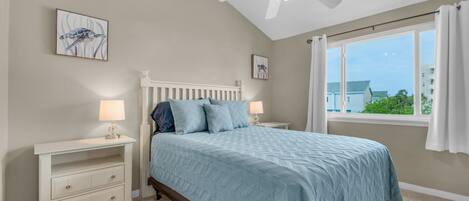 A bedroom with a bed, nightstands, a ceiling fan, and a window with a view of buildings and greenery.