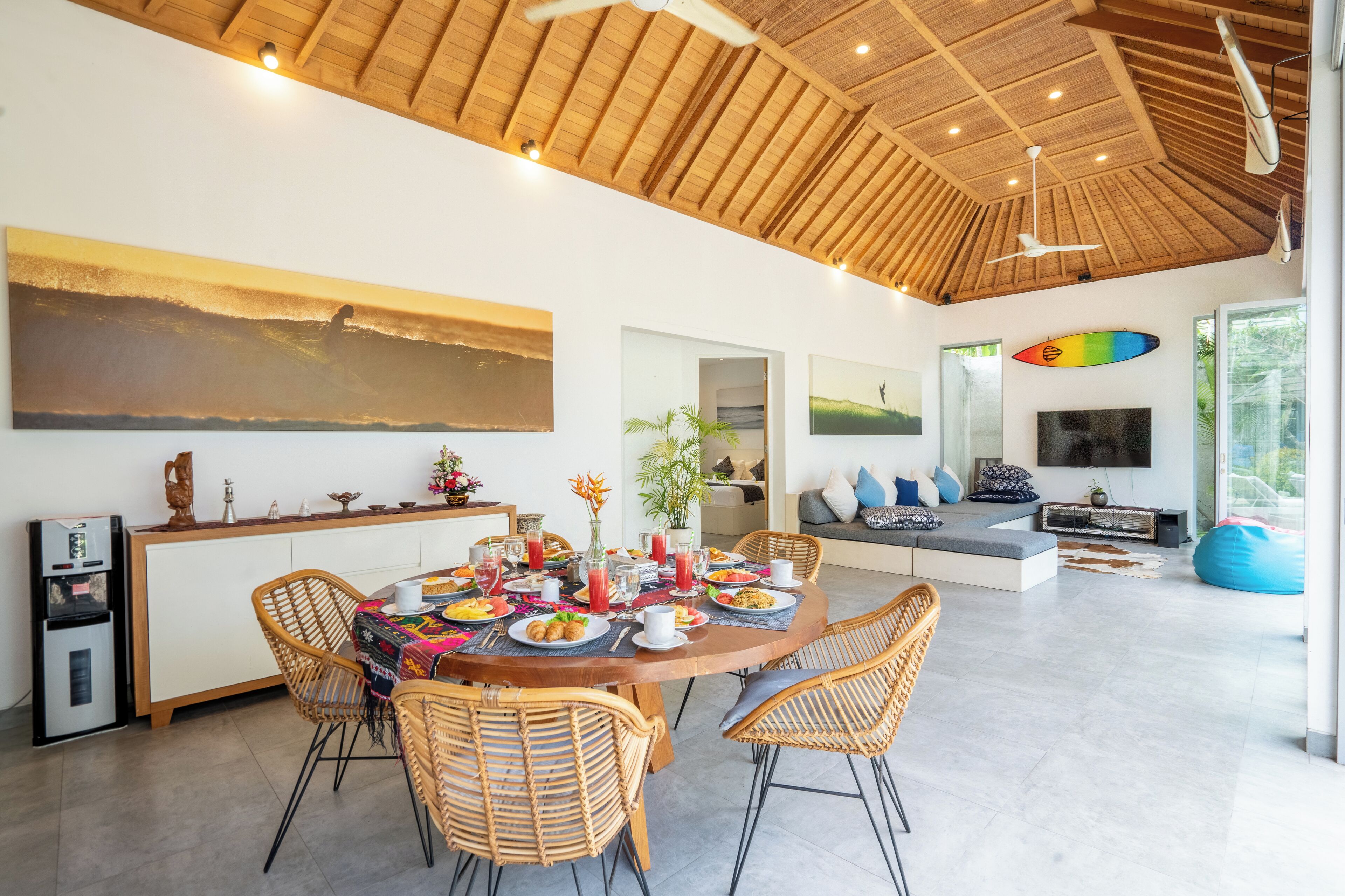 A spacious dining area with a wooden table and wicker chairs, a kitchen area with a refrigerator, and a living space with a sofa and television.
