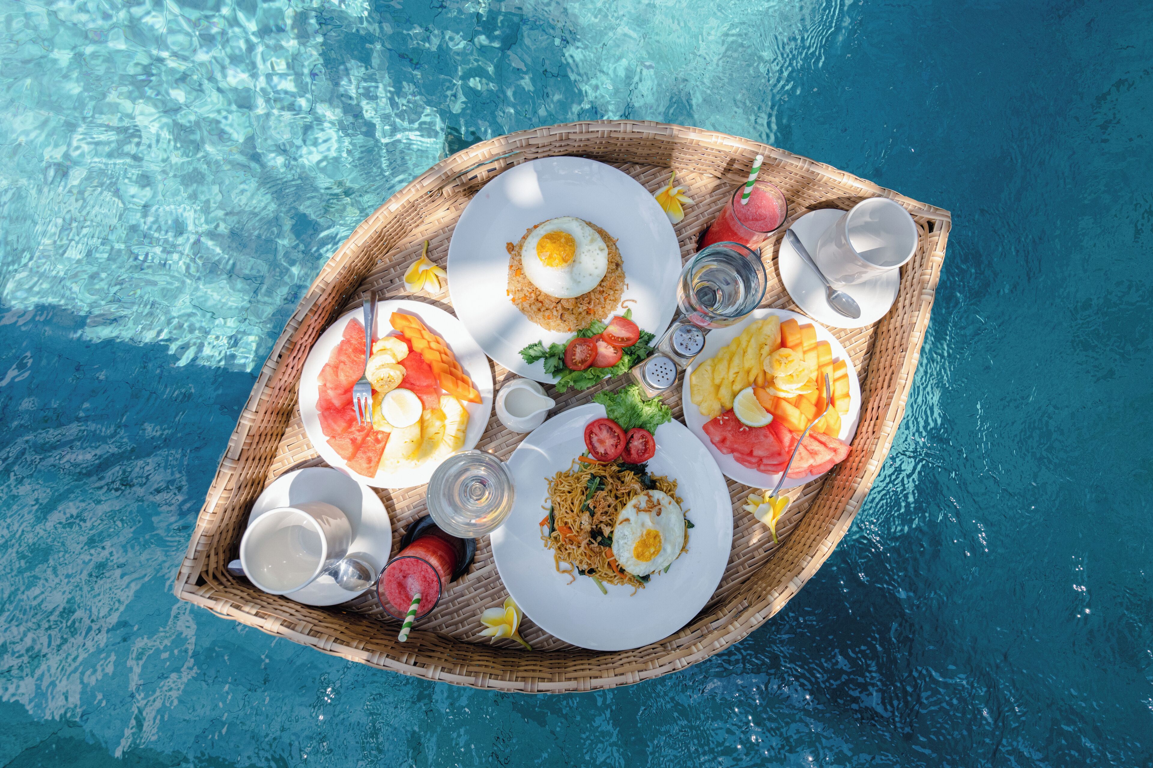 A wicker tray with breakfast items including eggs, noodles, and fruit, placed on a poolside.