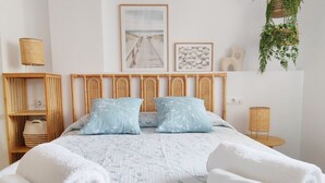 A neatly arranged bedroom with a bed, pillows, a bedside table, a lamp, a shelf with a basket, and wall art. - Appartment Sueños del Mar by DENIA COSTA (Denia)