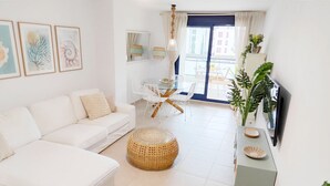 A modern living room with a white sofa, a round wicker ottoman, a dining table with white chairs, and a TV on a stand. - Appartment Sueños del Mar by DENIA COSTA (Denia)