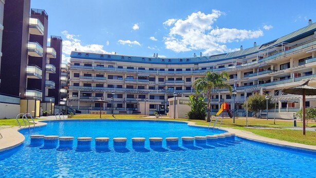 A swimming pool surrounded by a multi-story building with balconies, a playground, and a clear sky. - Appartment Sueños del Mar by DENIA COSTA (Denia)