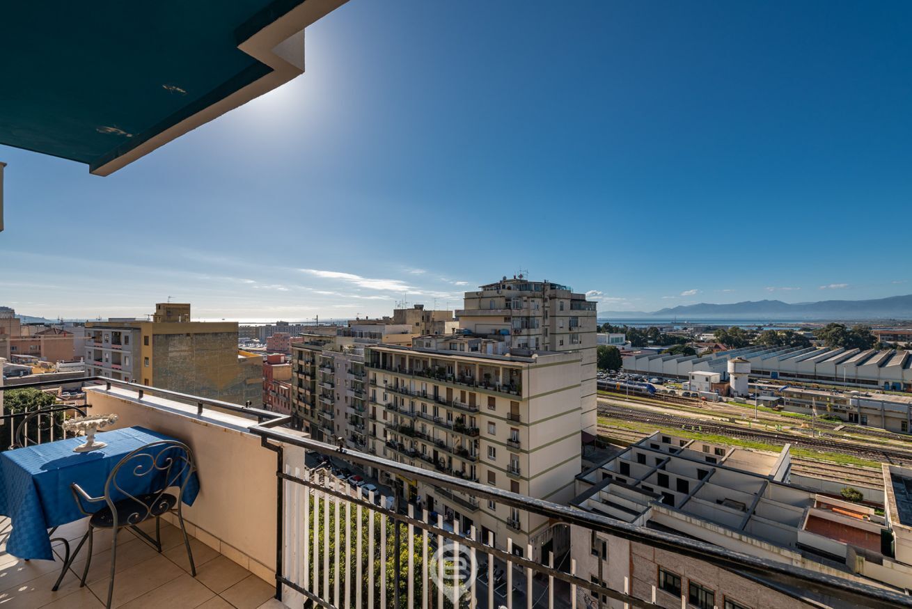 Un balcón con mesa y silla, con vistas a un paisaje urbano que incluye edificios y una vía de tren.