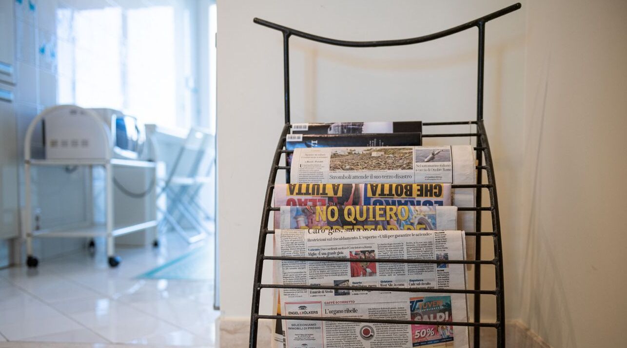 A metal rack with newspapers in a bright room with a white floor and a cart in the background.