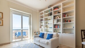 Un salon avec un canapé blanc, des coussins bleus, une grande bibliothèque et une vue à travers une porte vitrée.