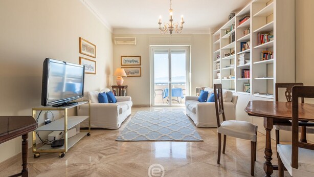 Un salon avec un téléviseur, des canapés, un lustre, une bibliothèque et une vue sur l’extérieur, visible à travers une porte vitrée.