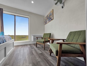 Une pièce dotée d’une grande fenêtre, de deux fauteuils verts, d’une table en bois et d’un tableau accroché au mur.