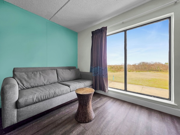 A modern living room with a grey sofa, a round wooden side table, and a large window with curtains. - Crystal Palace Resort (Port Bolivar)