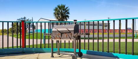 Una parrilla portátil con estructura metálica y ruedas, ubicada frente a un área cercada con cielo despejado y un edificio al fondo.