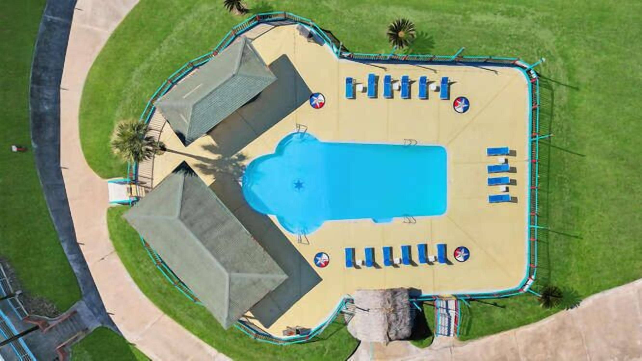 An aerial view of a resort with a swimming pool, sun loungers, and a fenced area.
