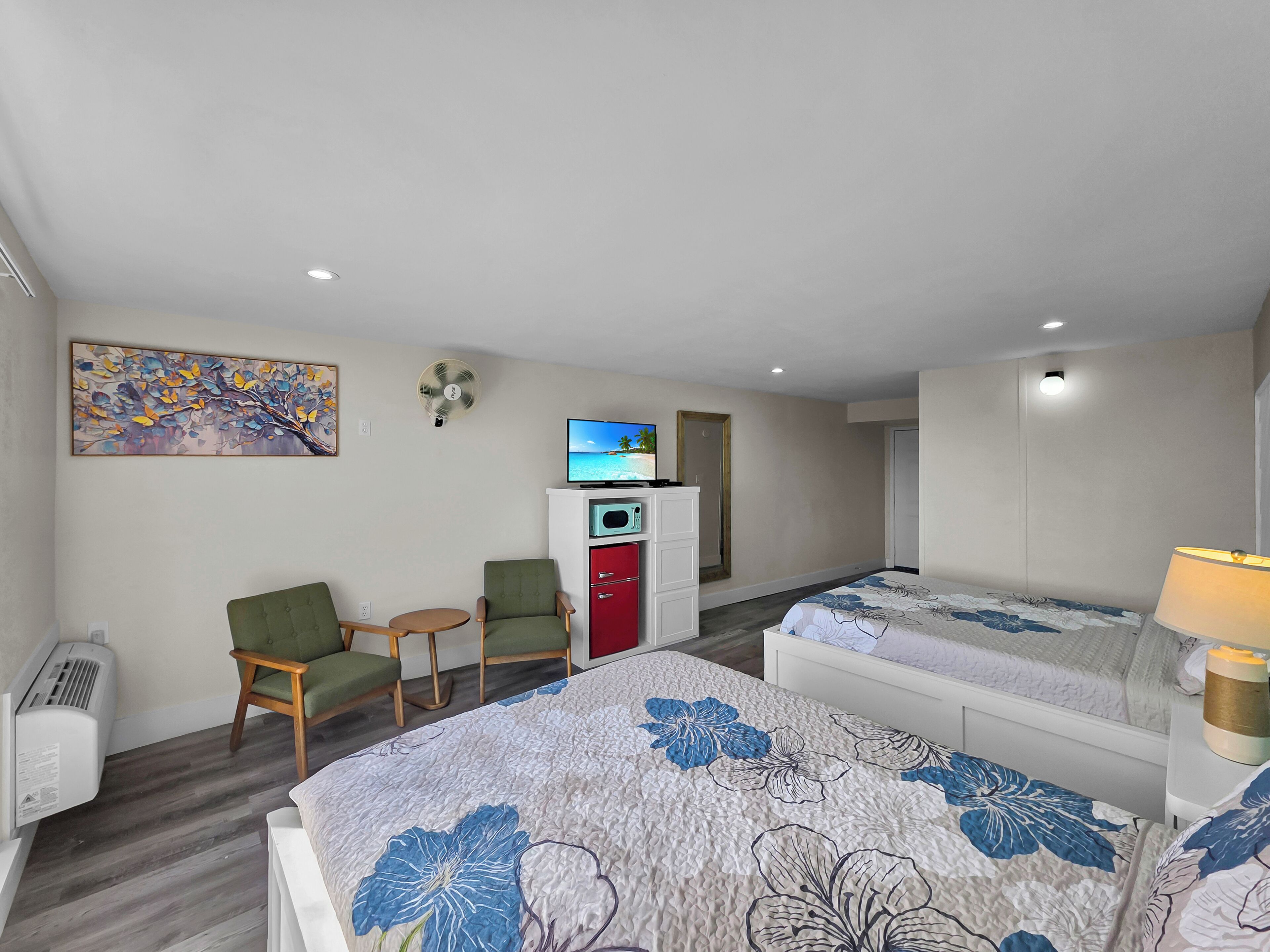 A hotel room with two beds, a desk with a TV, a chair, a small table, and a wall-mounted fan.