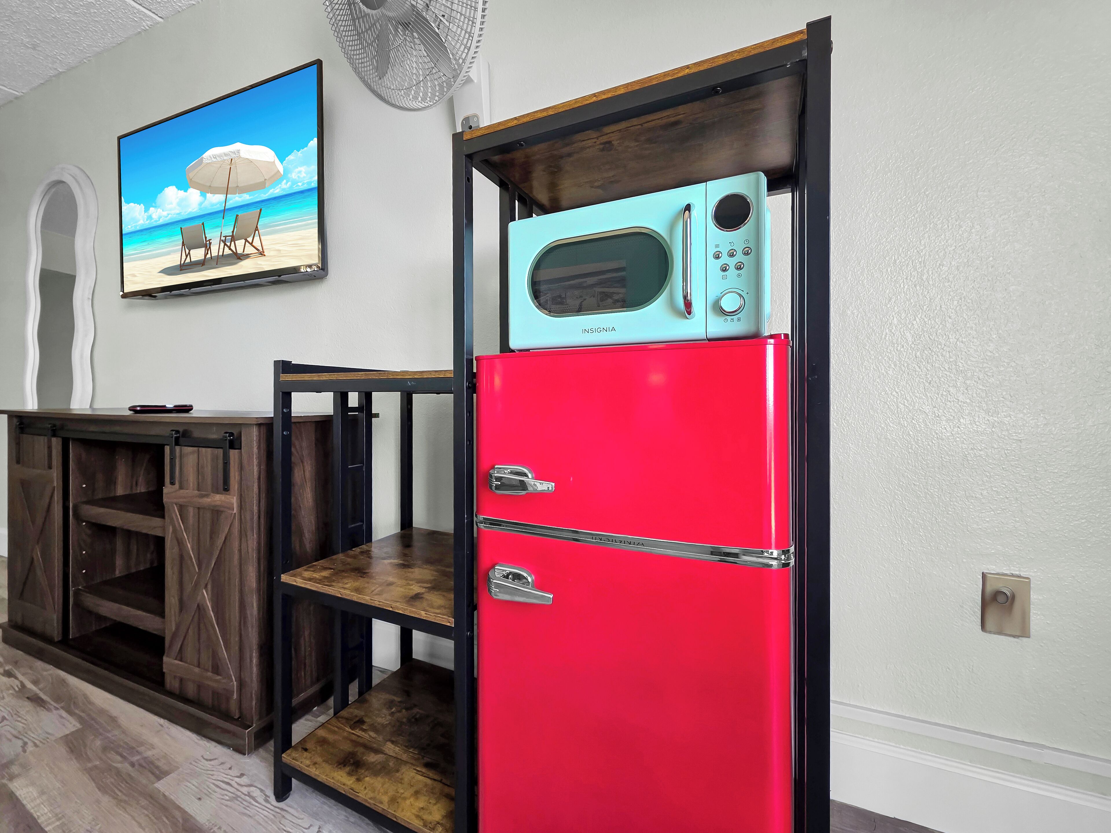 Una cocina con un refrigerador rojo, una mesa de madera y un ventilador fijado a la pared.
