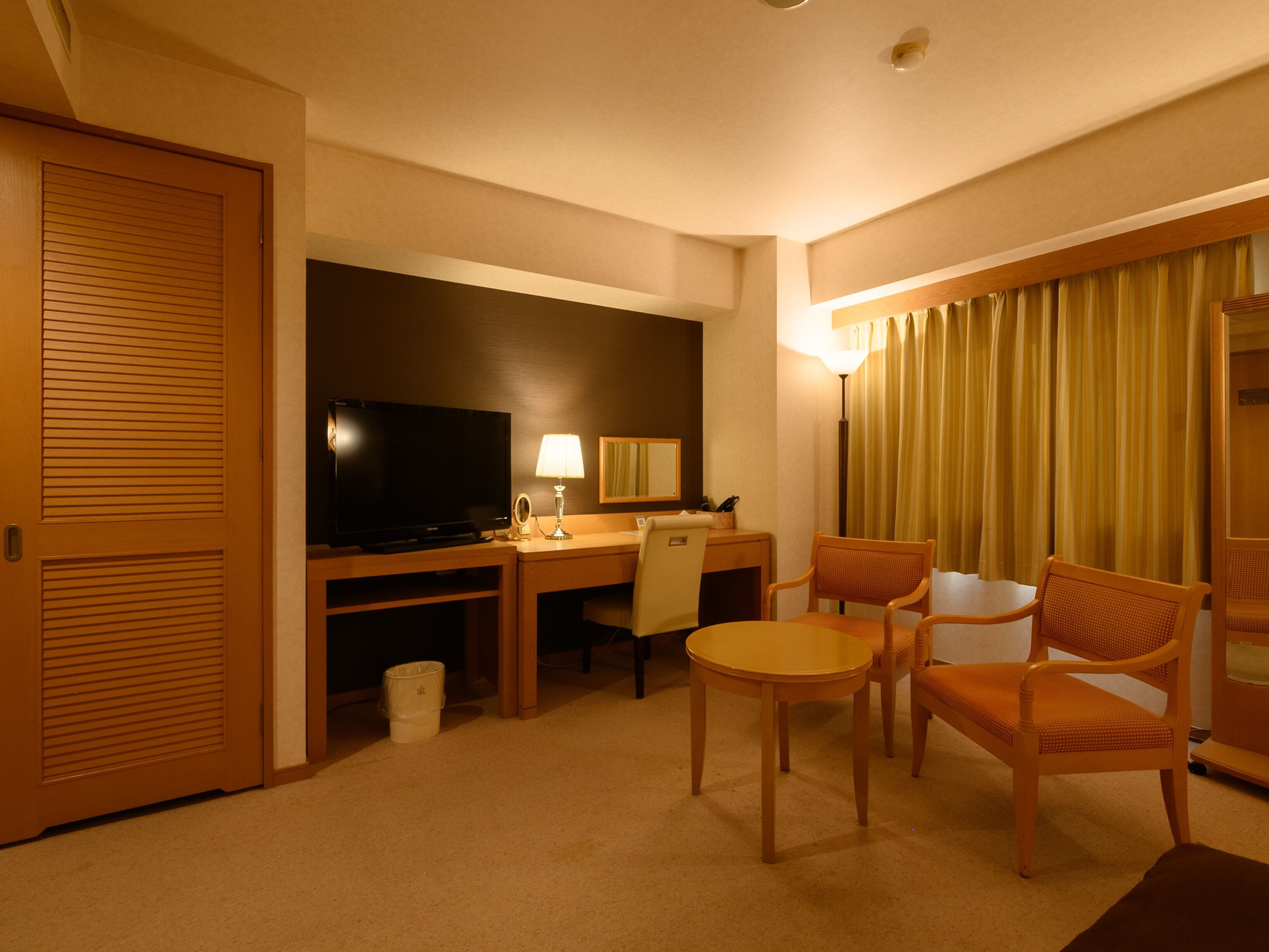 A hotel room with a desk, television, lamp, and two chairs.