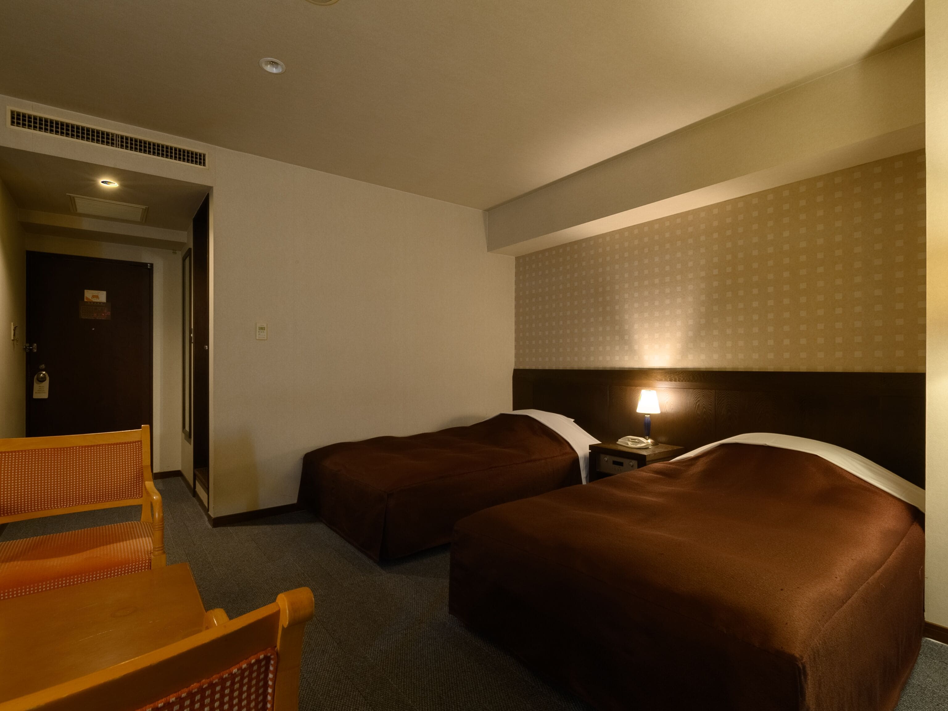 Ein Hotelzimmer mit zwei Einzelbetten, einem Holzstuhl, einem Nachttisch mit Lampe und einer Tür, die in einen anderen Raum führt.