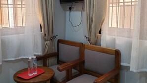 A corner room with a wooden chair and a small table, a TV mounted on the wall, and a window with curtains. - Hancol Hotel (Dodoma)