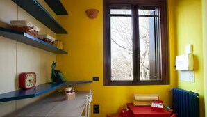 Una habitación con pared amarilla, una ventana, una mesa, sillas, un radiador y estantes con objetos.