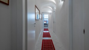 En gang med hvide vægge, flisegulv og røde tæpper, der fører til et rum med en seng og et vindue.