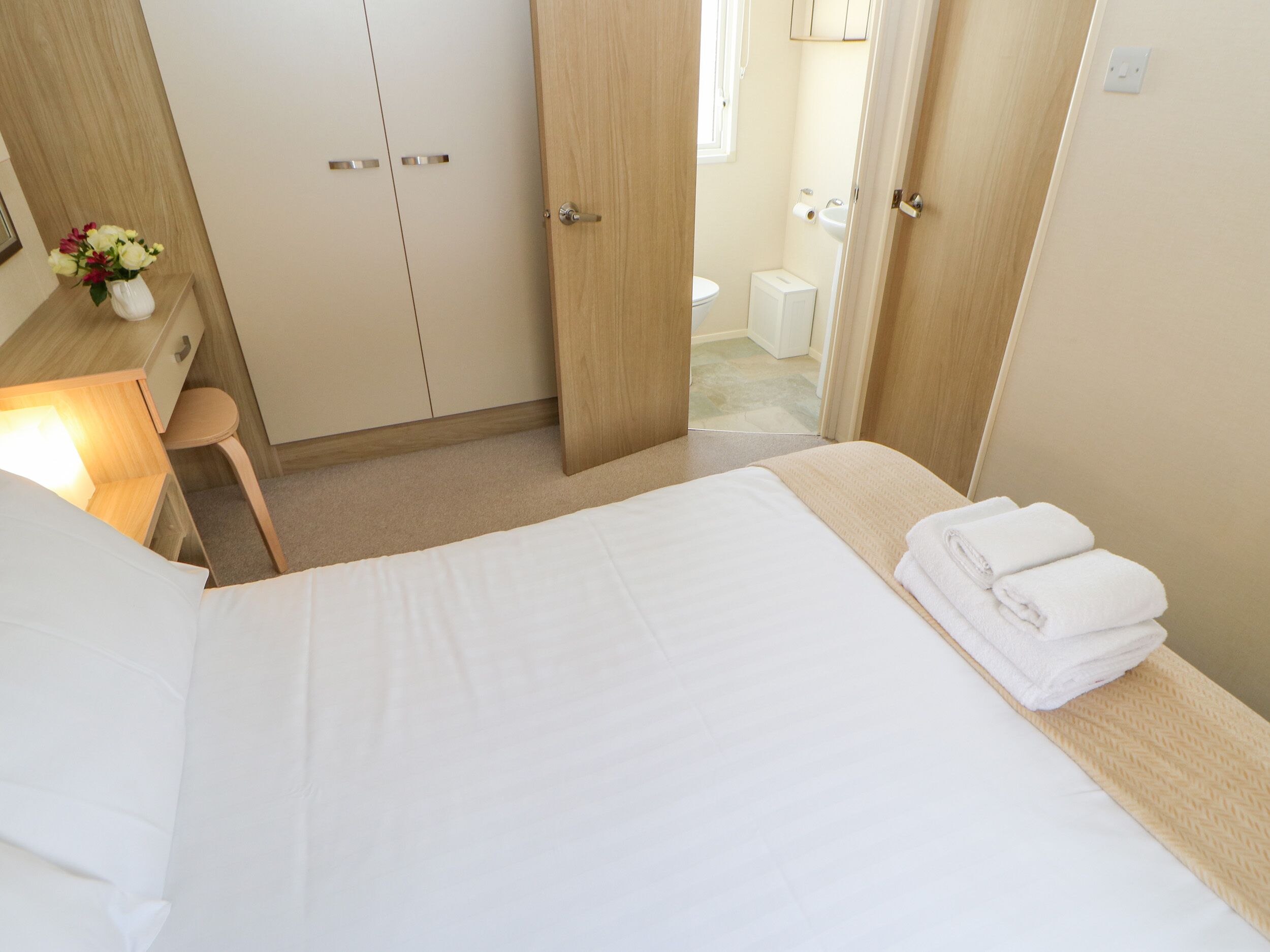 Habitación de hotel con cama, escritorio con lámpara, una pequeña mesa con un jarrón de flores y un baño visible a través de una puerta abierta.