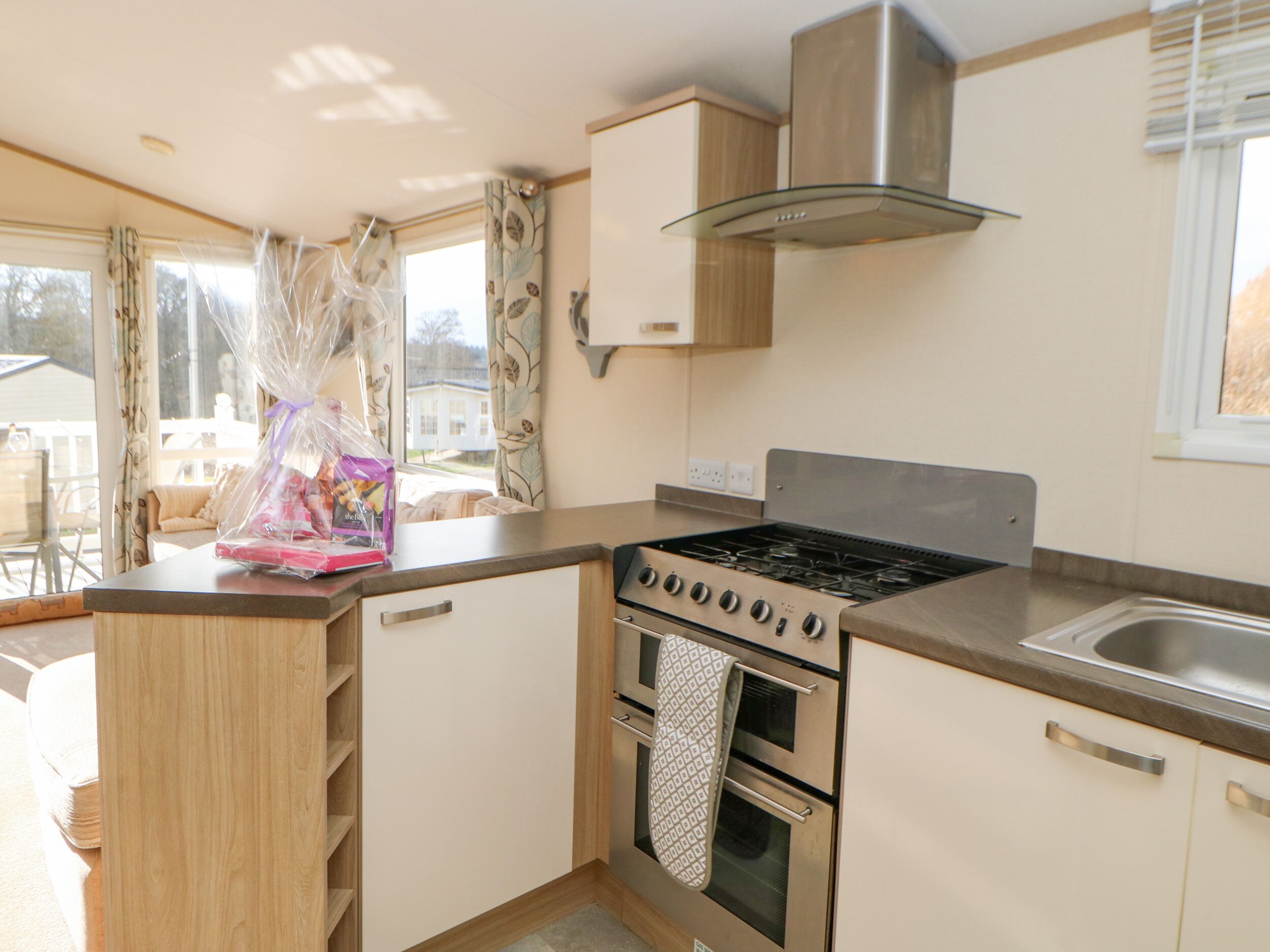 A compact kitchen with a gas stove, oven, and a stainless steel range hood. There is a window with curtains, and a small countertop with a bag of groceries.