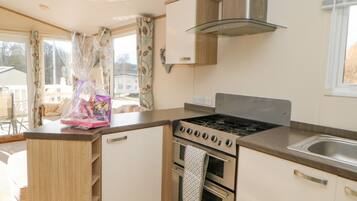 A compact kitchen with a gas stove, oven, and a stainless steel range hood. There is a window with curtains, and a small countertop with a bag of groceries.