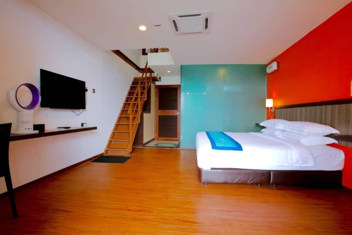 A hotel room with a bed, a TV mounted on the wall, a wooden staircase, and a red accent wall.