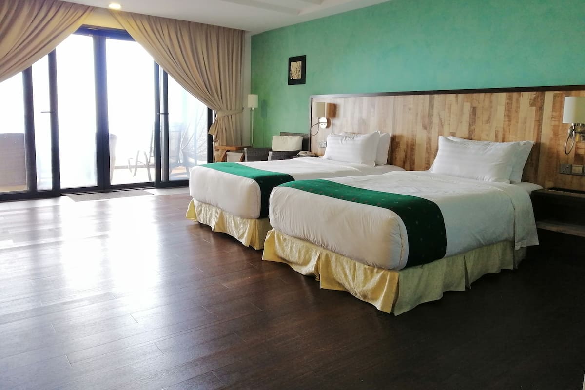 A hotel room with two beds, a wooden headboard, a green wall, and a large window with curtains.