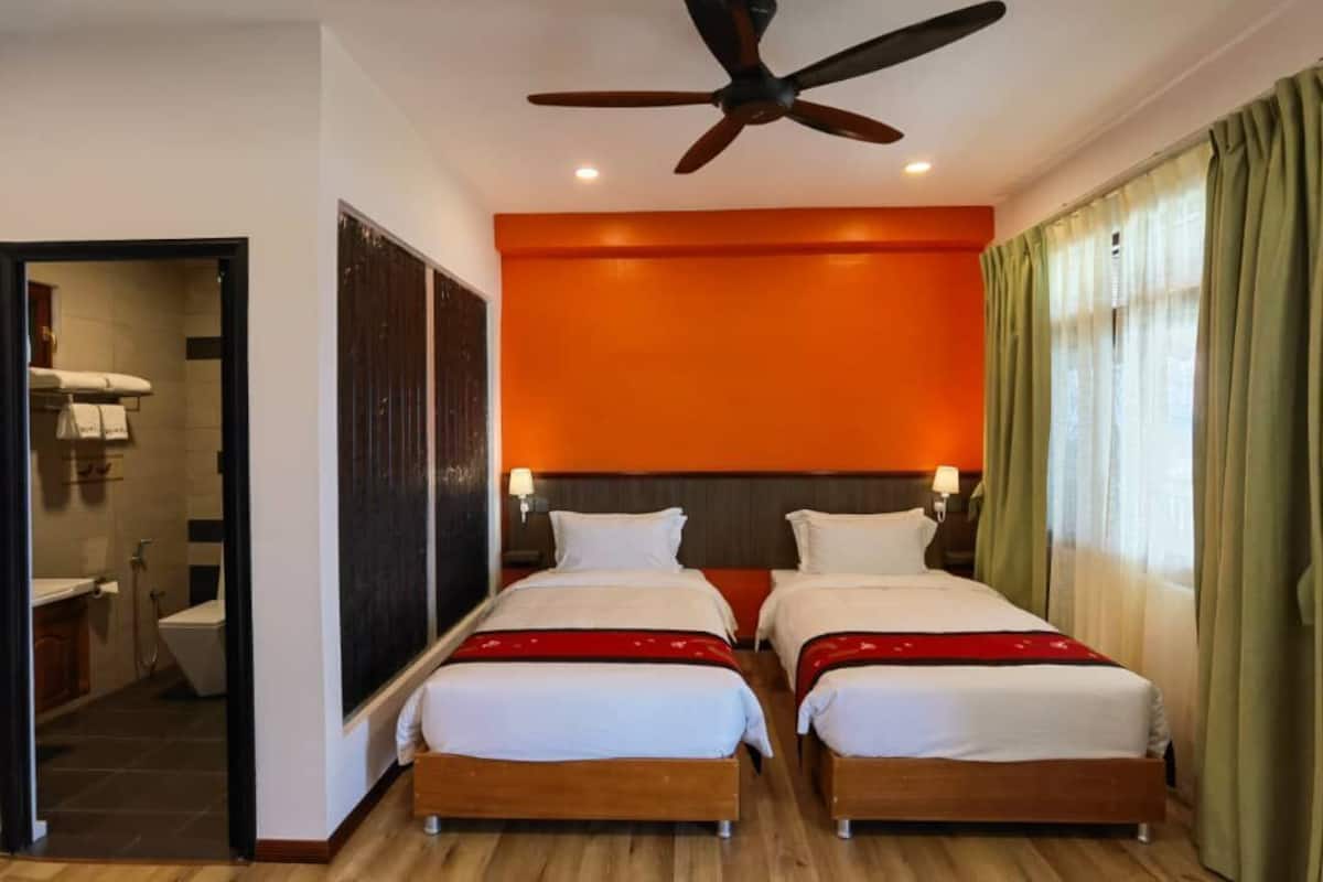 A hotel room with two beds, a ceiling fan, an orange accent wall, and a view of the city through the window.