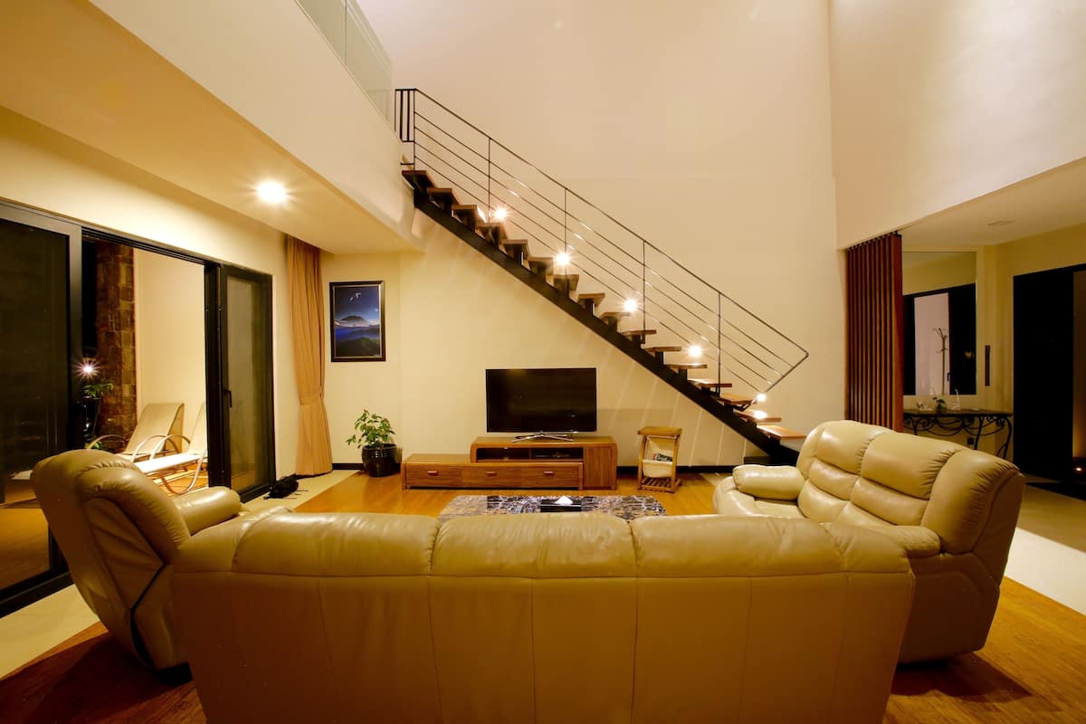 A modern living room with a staircase, leather sofa, entertainment unit, and a flat-screen TV.