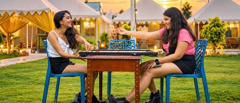 Two individuals playing a board game outdoors with a table and chairs, tents in the background, and a grassy area.