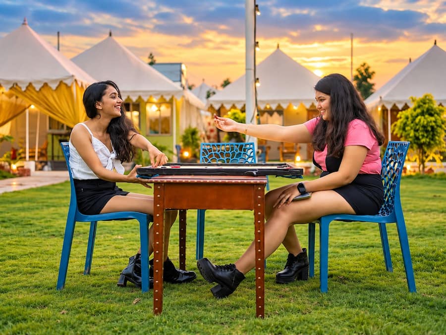 Two individuals playing a board game outdoors with a table and chairs, tents in the background, and a grassy area.
