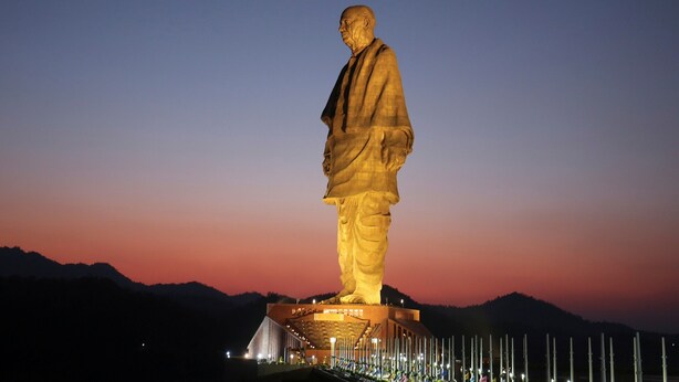 Une grande statue illuminée représentant un homme debout sur une plateforme, avec en arrière-plan des montagnes et un ciel crépusculaire.