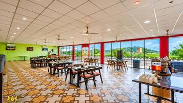 A spacious dining area with multiple tables and chairs, large windows offering a view of the outdoors, and a ceiling with recessed lighting and fans.