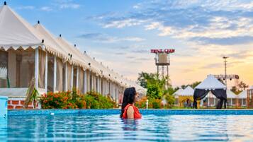 Una piscina con una persona en el agua, una fila de tiendas de campaña blancas y una torre con un letrero al fondo.