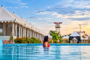 Une piscine avec une personne dans l’eau, une rangée de tentes blanches et une tour avec une enseigne en arrière-plan.