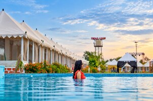 A swimming pool with a person in the water, a row of white tents, and a tower with a sign in the background.